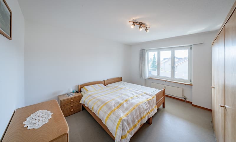 Bedroom with a double bed, wooden nightstands, a doily on a table, a picture frame on the wall, a ceiling lamp, and a window with white curtains.