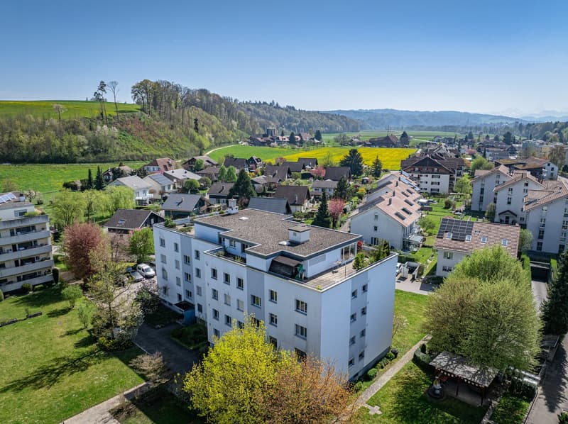 Aerial view of a white apartment building with multiple windows, surrounded by a green landscape with trees and houses.