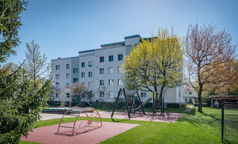 Modern apartment building with multiple windows, surrounded by trees and a playground.