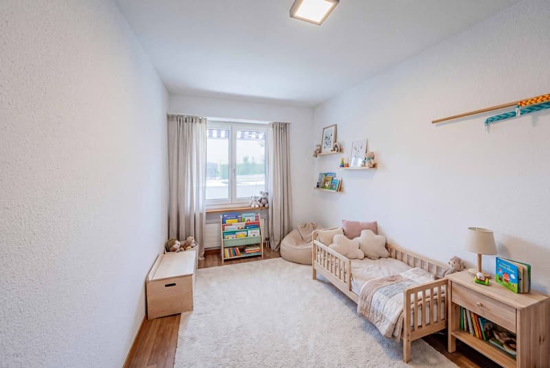 Child's bedroom with white walls, wooden floor, white rug, bed, shelves, lamp, storage box, bean bag, toys, books, picture frames, window with curtains