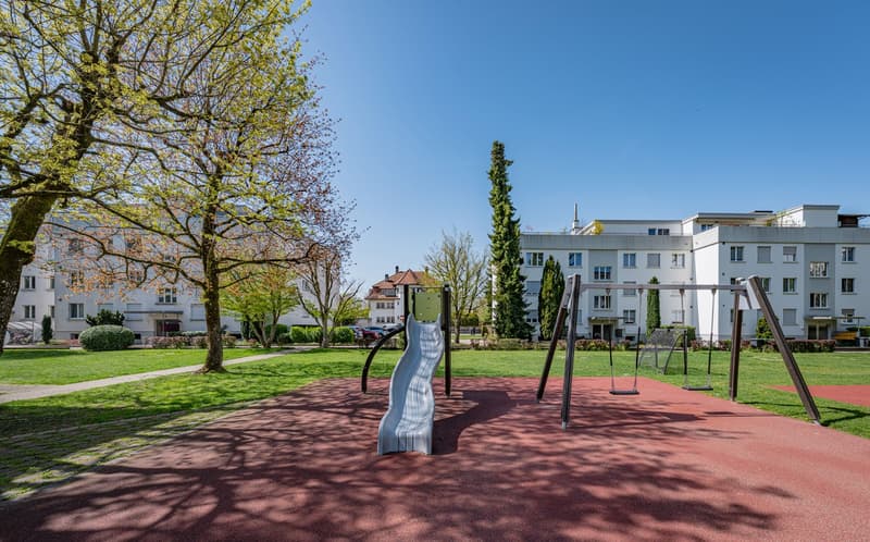 Playground with slide and swings, surrounded by trees and grass, buildings in the background