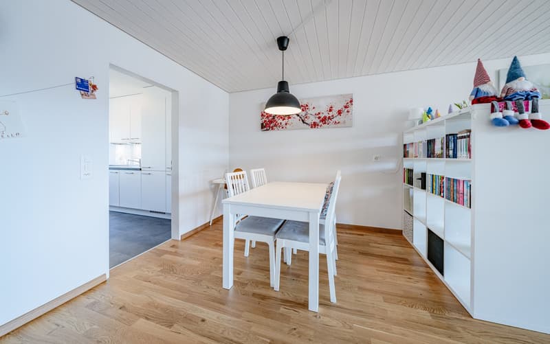 Dining room with white table and chairs, wooden floor, white walls, white ceiling, white bookshelf, white kitchen cabinets, hanging lamp, picture frame, gnomes on bookshelf