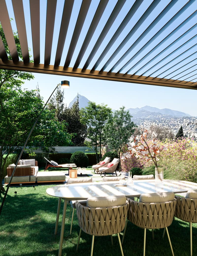 A spacious outdoor garden area with a rectangular table and cushioned chairs. There is a couch with pillows on the left side, a lamp on a stand, and a decorative plant in a vase. An outdoor canopy provides shade over the seating area, and there is a view