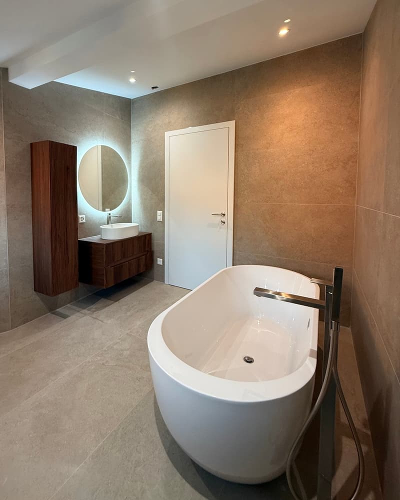 contemporary bathroom with a white bathtub, large sink, mirror, wooden cabinet, and beige tiles