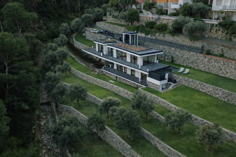 Modern house, multiple levels, large windows, terrace, surrounded by stone walls, greenery, olive trees