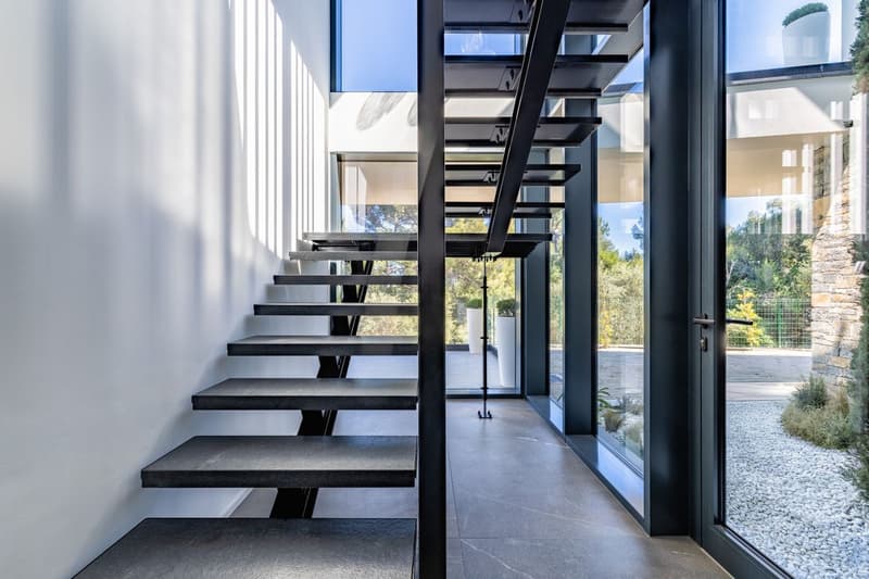Modern staircase with black steps and metal railings, large glass windows, white walls, and a view of the outside landscape.