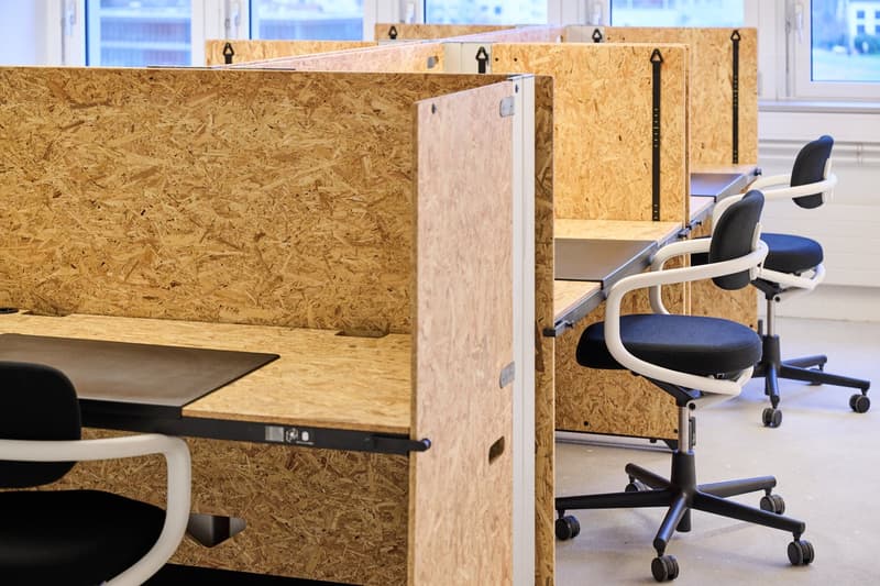 Open office layout with wooden dividers, desks with metal bases, black and white office chairs, large windows