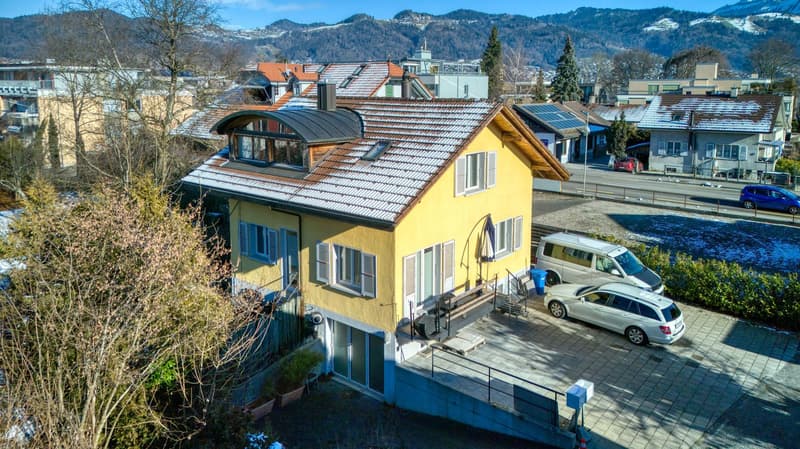 Yellow 2 story house, brown roof, solar panels on another house, 2 cars parked in front, garage entrance