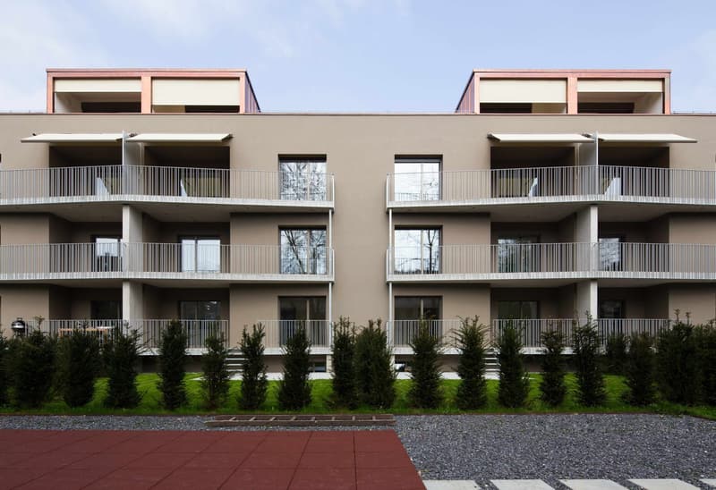 Three-story apartment building with balconies on each floor, brick walls, glass windows, and a red gravel path.