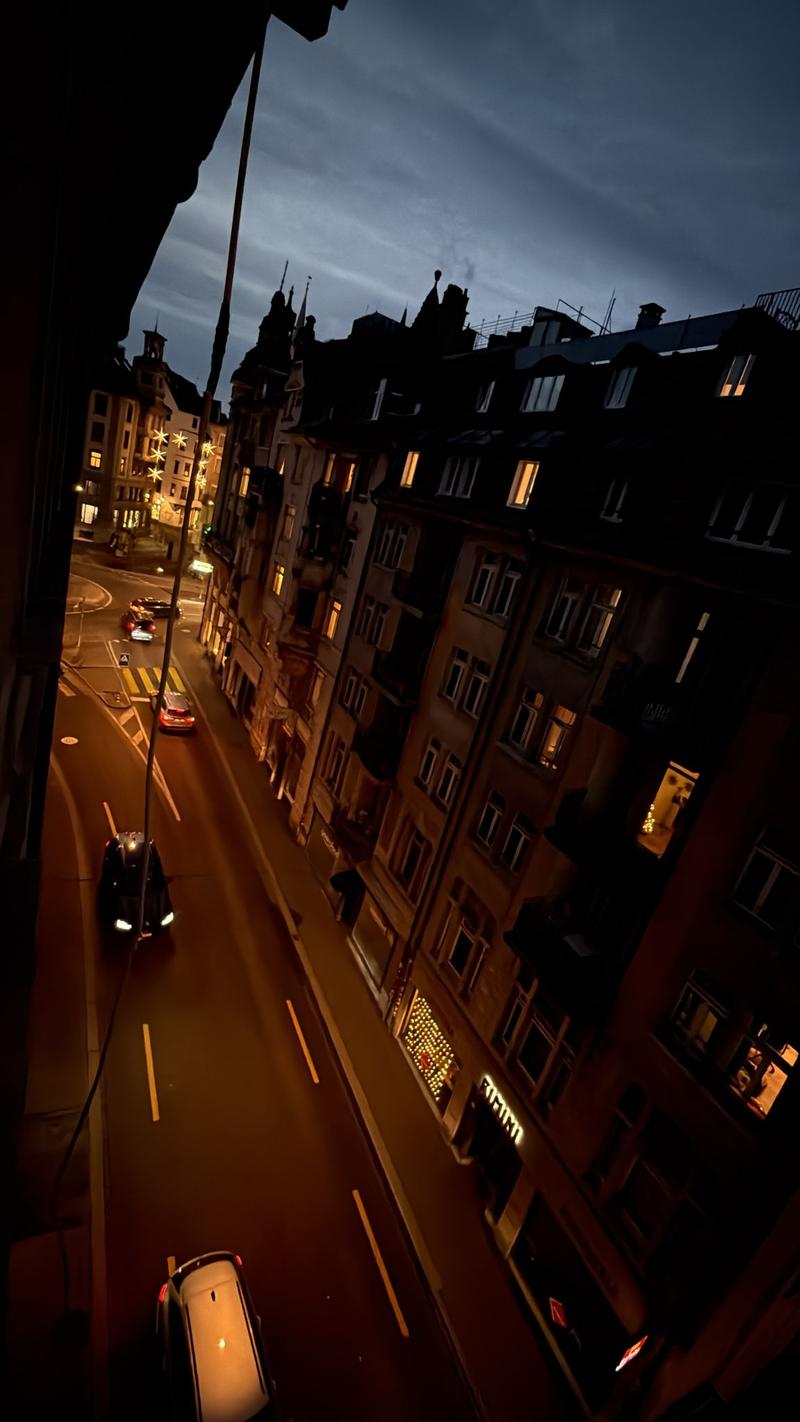 a street scene at night, with multiple multi-story buildings on either side of the road, several cars driving down the road, and a view of the city skyline