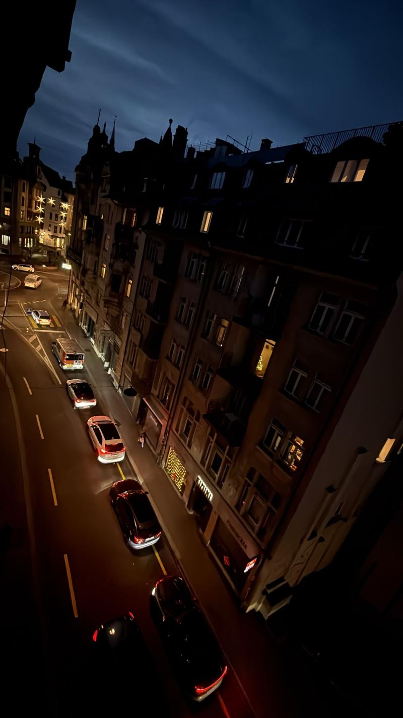 An exterior view of a building at night, showing multiple cars driving on the road, street lights, and buildings with windows and some illuminated.