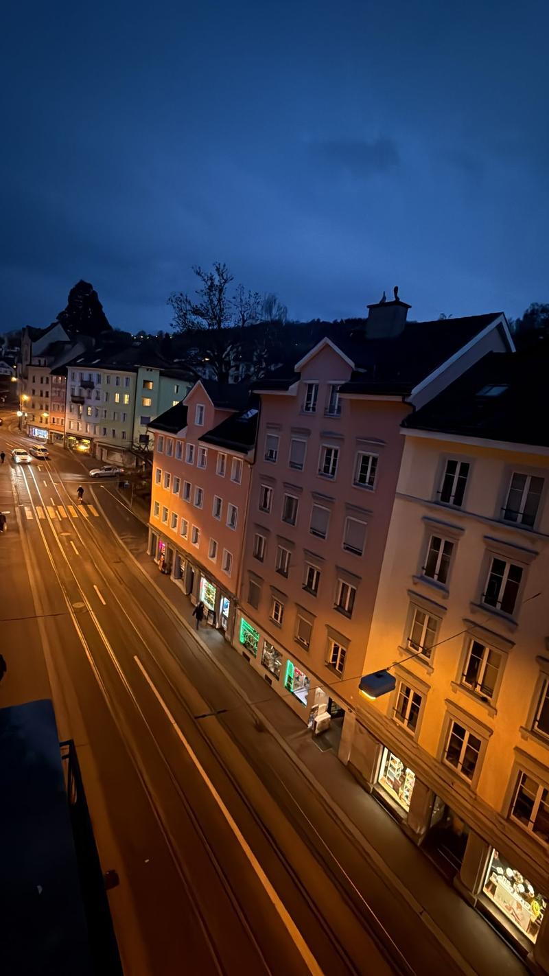 City street at night, with buildings, vehicles, streetlights, pedestrians, and greenery.