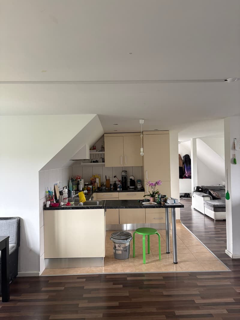 Open-plan kitchen with dining table, white kitchen cabinets, black granite countertops, green stool, waste bin, flower vase, kitchen tools, microwave, coffee machine, sink with faucet