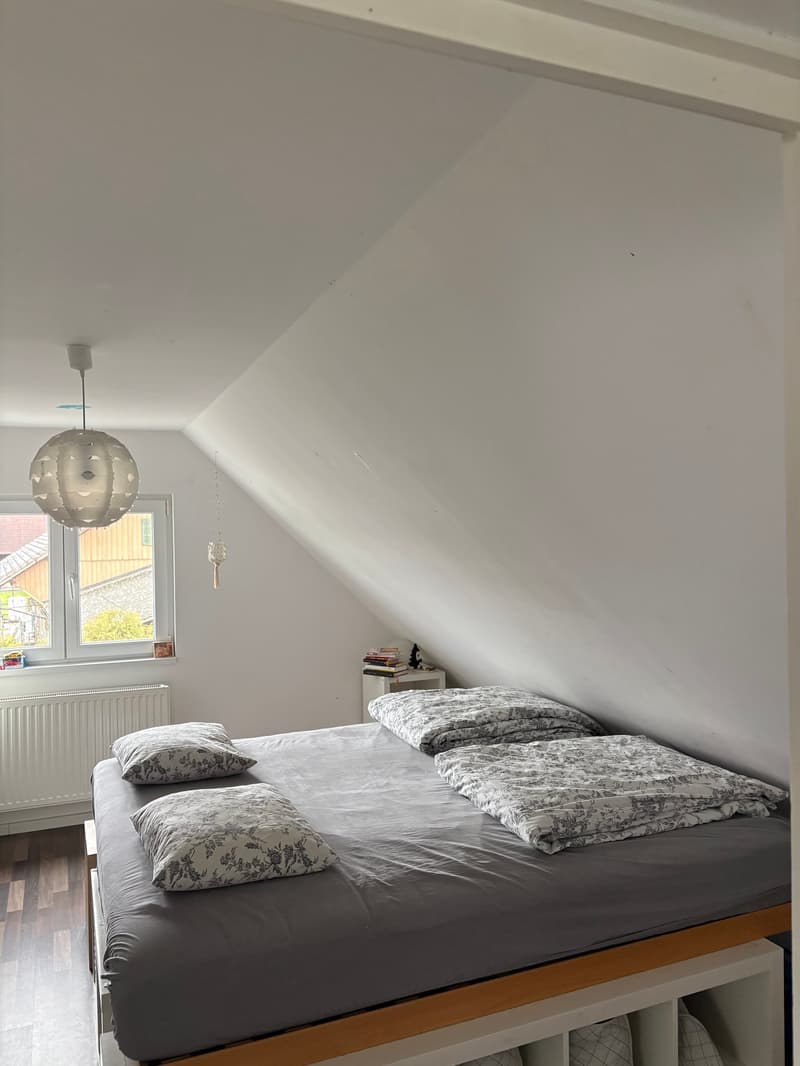 Bed, pillows, blankets, white walls, white wooden floor, lamp, nightstand with books, radiator, window with view