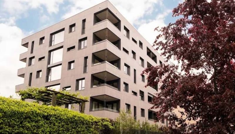 Modern apartment building with several balconies, gray exterior, many glass windows, hedges and trees