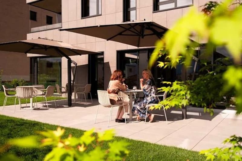 An outdoor seating area with tables and chairs under umbrellas, women sitting, building in the background, grass, trees