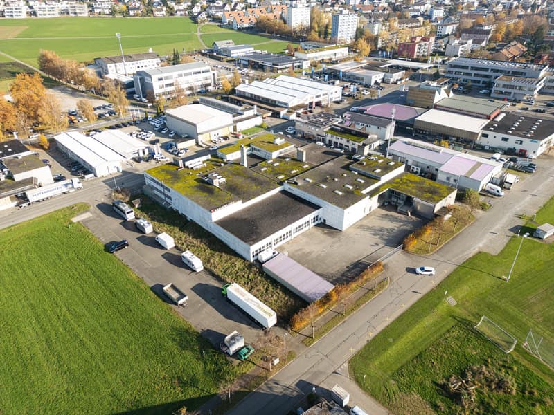 Industrial buildings with green roofs, parking lot with cars, white trucks, road, large field, and distant city buildings