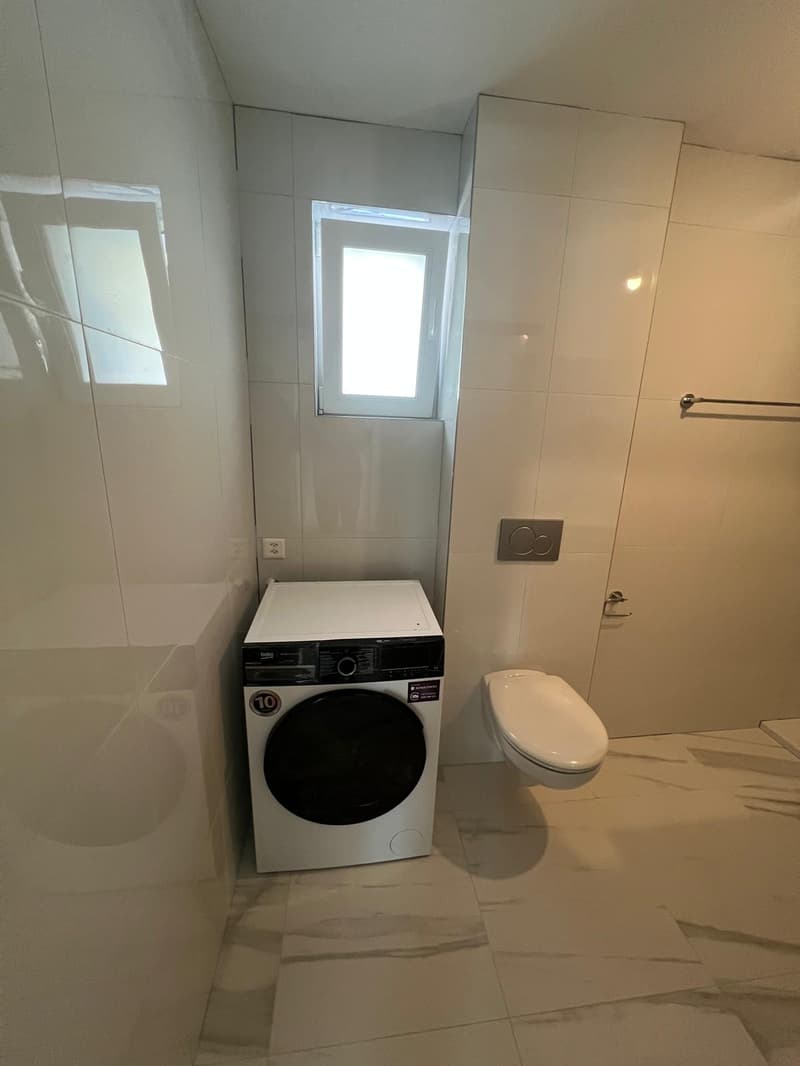 Small bathroom with white tiles, washing machine, toilet, and a window