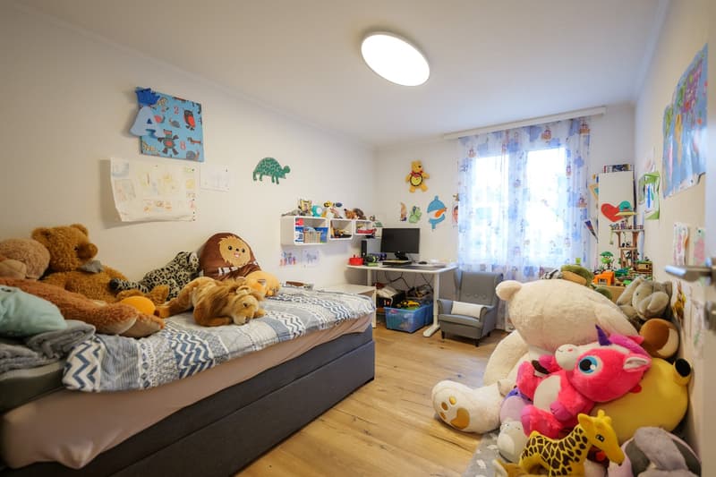 Children's bedroom with bed, numerous stuffed animals, small desk with monitor, grey armchair, and colorful drawings on the wall