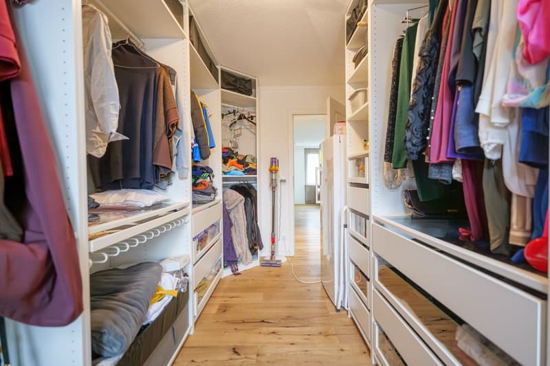 Spacious walk-in closet with wooden floors, white shelves, various clothing items, vacuum cleaner, white refrigerator