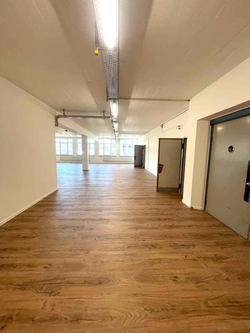 Empty room with wooden floors, white walls, fluorescent ceiling lights, windows, doors.