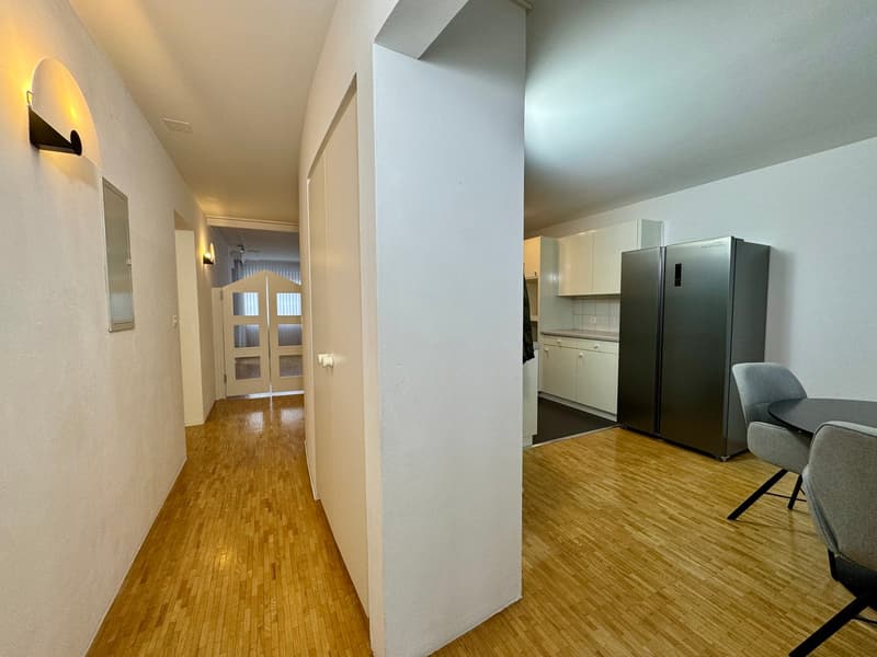 The hallway features wooden flooring, white walls, a modern light fixture on the left wall, and an open door leading to another room with a glass window. On the right side, there is a kitchen area equipped with white cabinets, a stainless steel refrigerat
