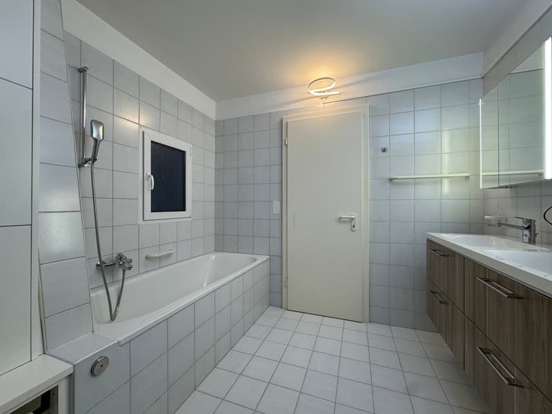 White tiled bathroom, bathtub with shower, single sink vanity, mirror, wall-mounted towel rack