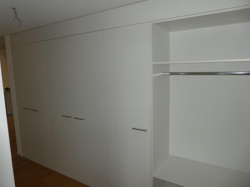 A white built-in wardrobe with sliding doors and shelves, mounted on the wall, with a wooden floor.