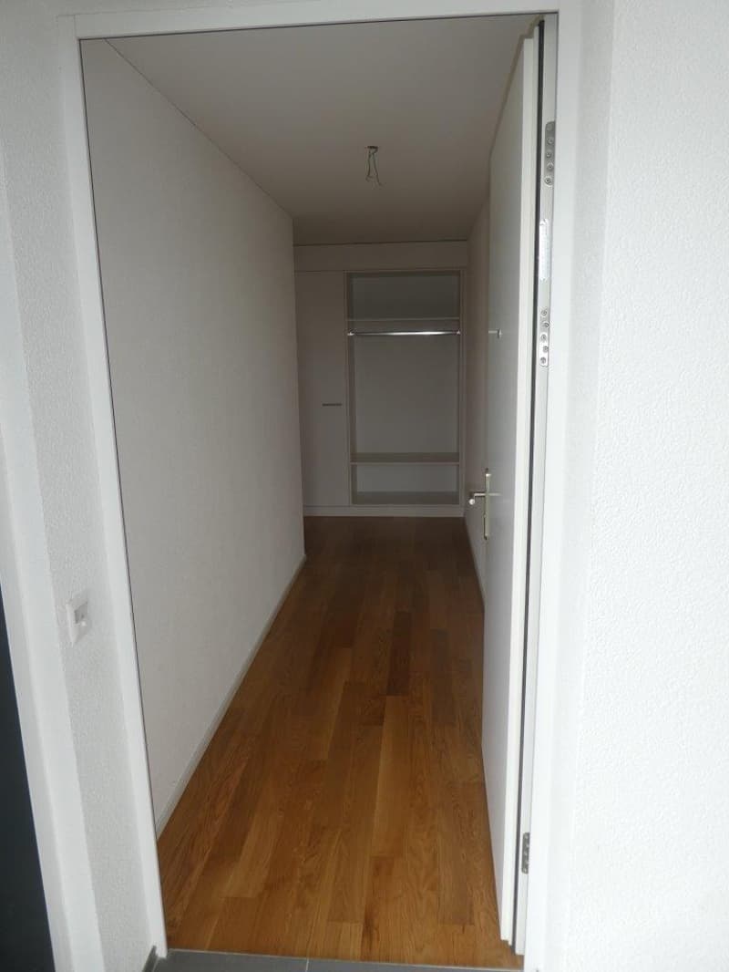 Empty hallway with wooden floor, white walls, open door, and a closet with shelves