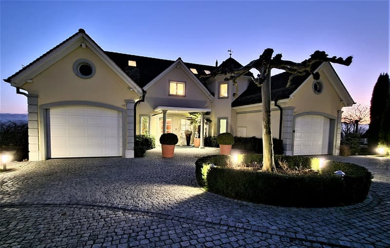 2 story house, beige exterior, black roof, 2 garages, paved driveway, hedges, outdoor lighting
