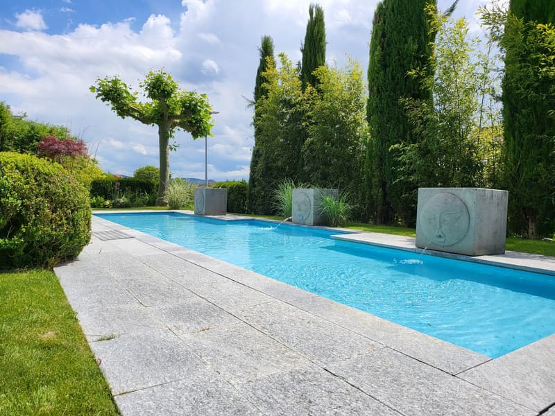 Outdoor swimming pool, concrete pool deck, two concrete sculptures, green lawn, bushes, trees
