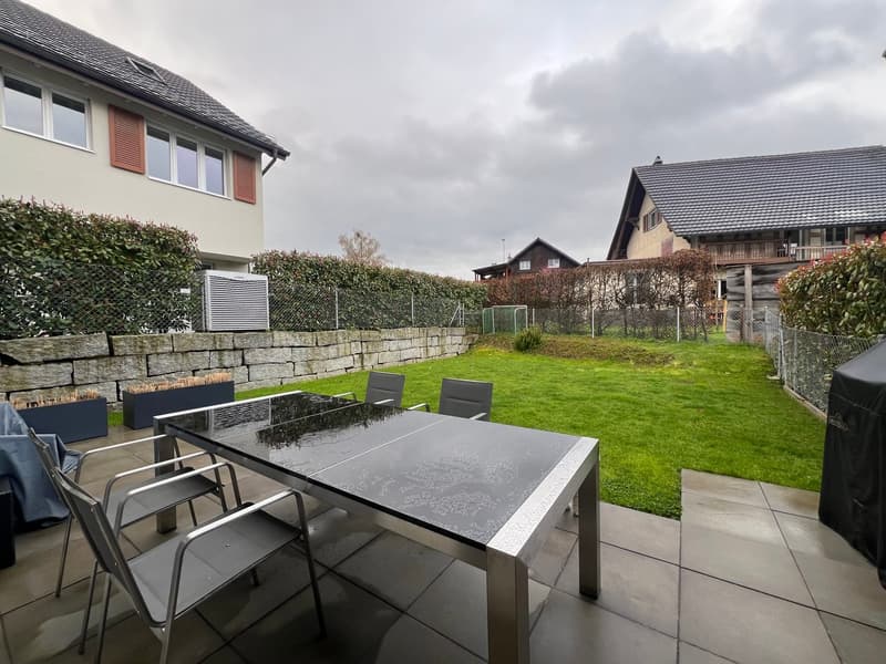 Outdoor dining table with chairs, covered by a table cloth, stone wall, green lawn, plants, houses in the background
