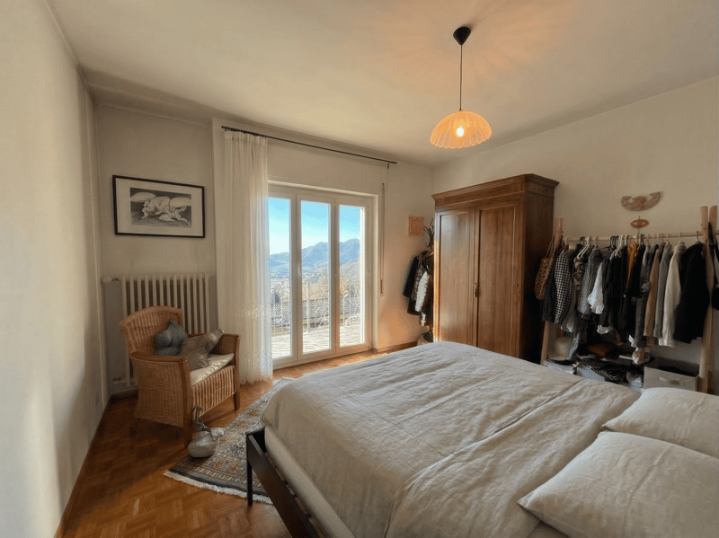 bedroom with wooden floor, large windows, bed, wooden chair, wooden cabinet, radiator, picture frame, rug, lamp