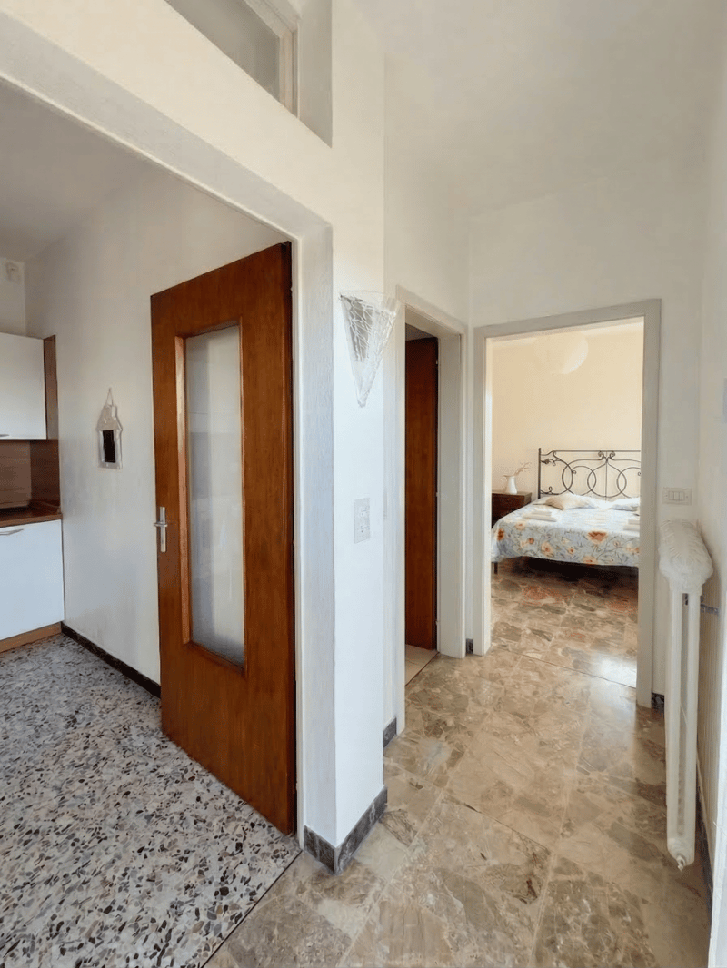 Entrance hall with wooden door, white walls, marble floor, kitchen area with cabinets, bedroom with bed and nightstand