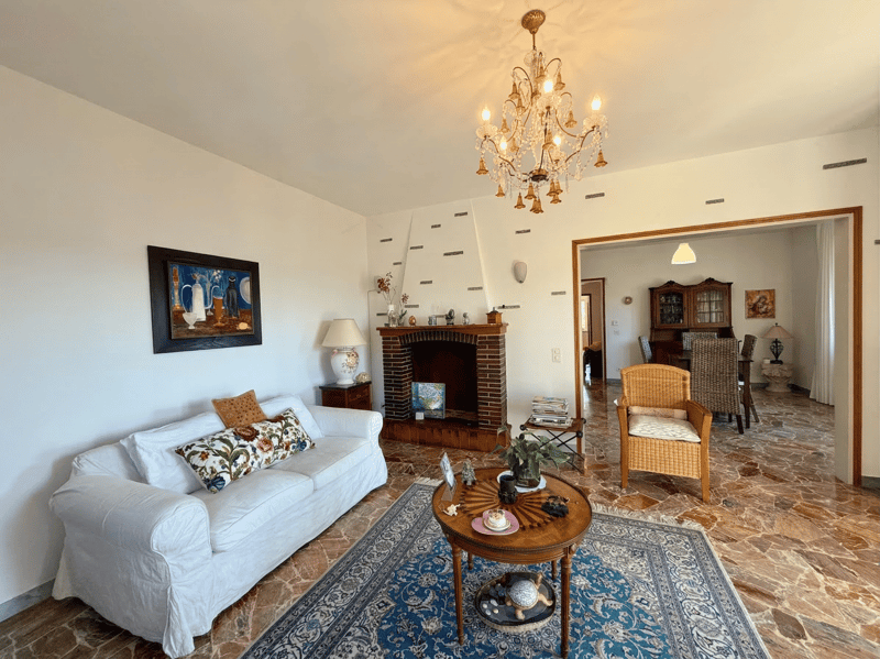 white couch, fireplace, chandelier, wooden coffee table, rug, side table with lamp, painting, wooden chair