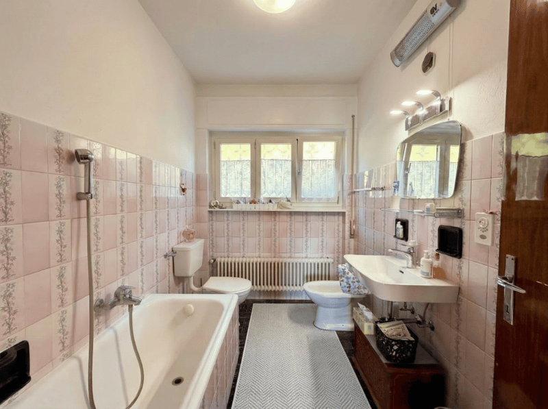 Bathroom with bathtub, toilet, bidet, sink, mirror, radiator, tiled walls, window, shower head, basket, and a rug.