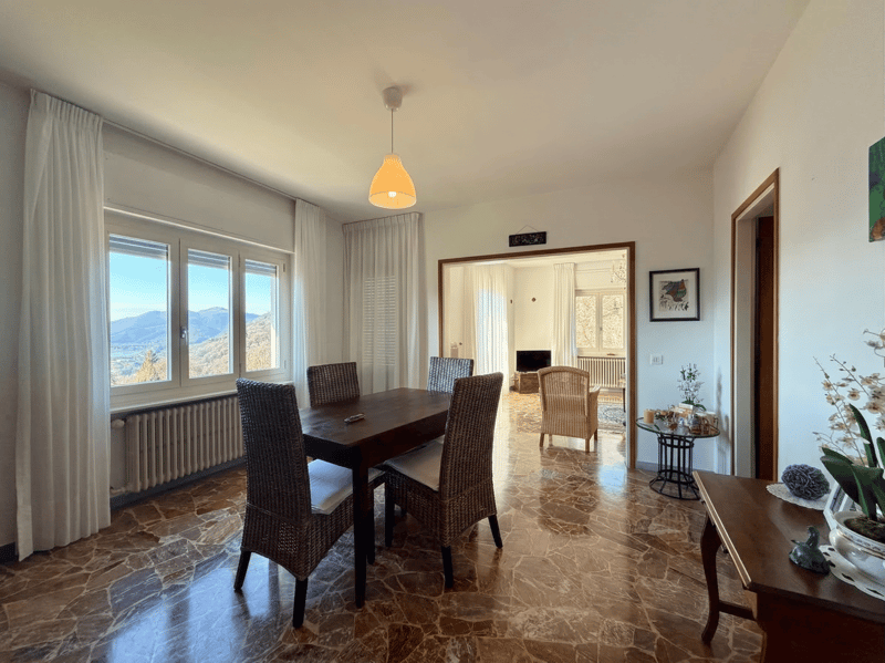 Dining room with wooden table, wicker chairs, white curtains, radiator, lamp, picture frame, side table, and a view of the mountains