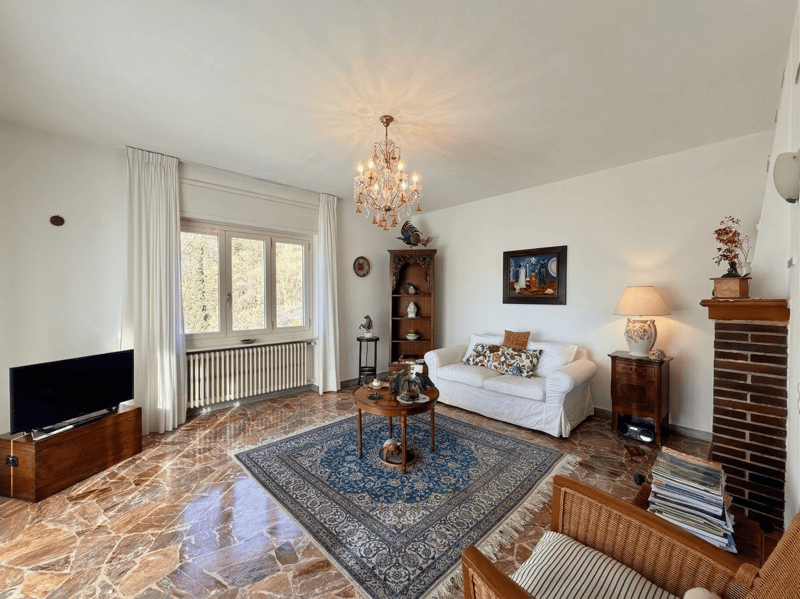 white walls, chandelier, fireplace, white couch, coffee table, rug, TV, wooden furniture, lamp, picture frame, radiator