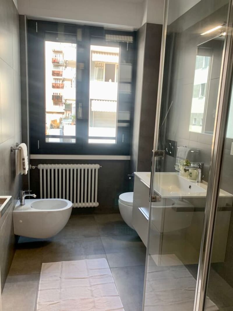 Bathroom with white fixtures, glass shower enclosure, white sink, white toilet, white bidet, white towel, white towel rack, white radiator, white floor, white walls, window with view