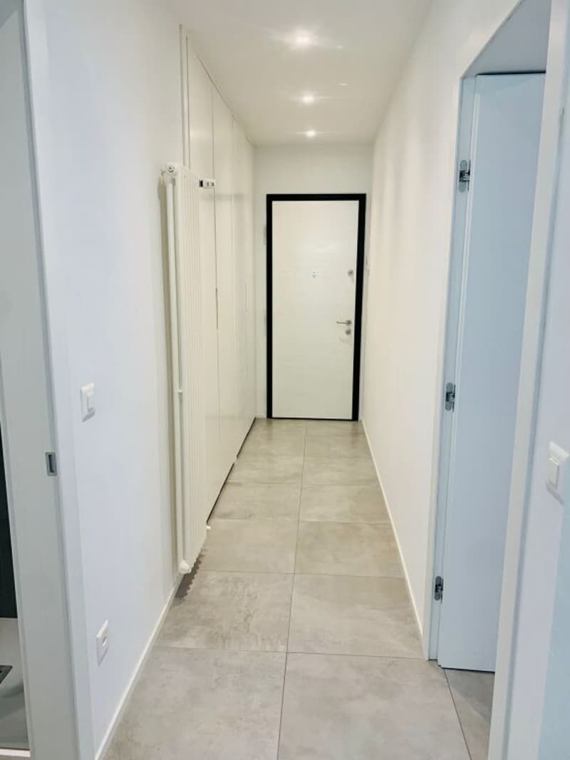 Empty hallway with white walls, tiled floor, white doors, and a white radiator on the left wall.