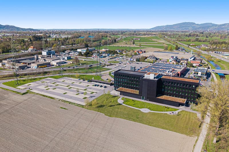 A modern building complex surrounded by green fields, featuring a large parking area, a landscaped pathway leading to the entrance, and large windows