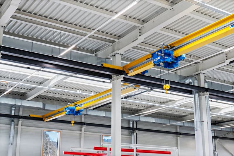 Large warehouse with white and grey walls, white ceiling with hanging lights, yellow and blue overhead cranes.