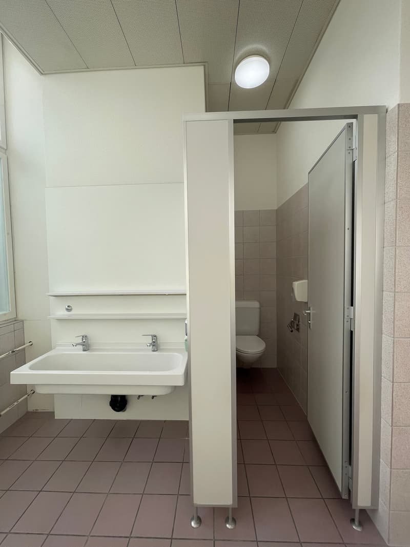 Bathroom with white sink, two faucets, a toilet, and a small window. Tiled floor, tile partitions, and a ceiling-mounted circular light fixture.