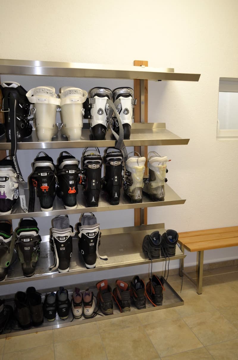 Many boots are stored on a steel shelf, wooden bench at the side of the shelf, tile flooring.
