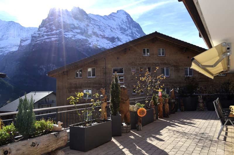 wooden house, balcony with railing, wooden statues, outdoor seating, garden with plants, mountainous background