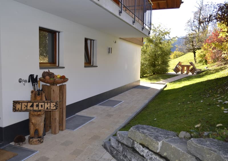House entrance, welcome sign, umbrella stand, tiled walkway, balcony, windows, garden with trees, pathway, rocks