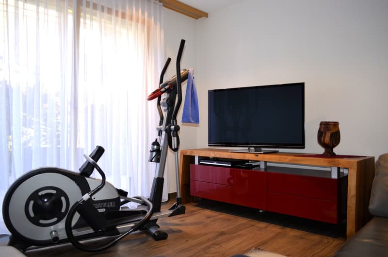 Room with wooden floor, two exercise bikes, wooden entertainment unit with TV, and red drawers.