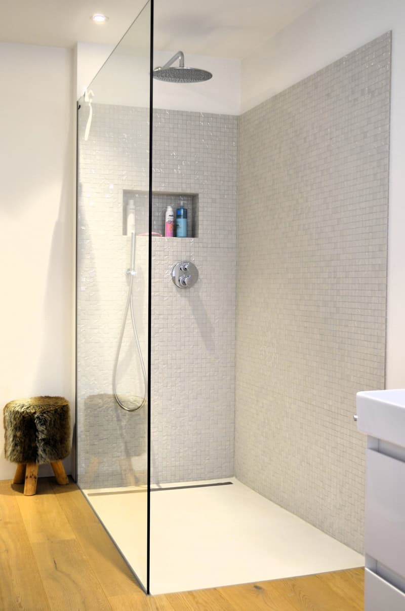 Modern shower area with glass enclosure, tiled walls, wooden floor, and a shelf with toiletries.