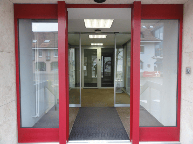 An exterior entrance to a building with sliding glass doors, red frames, and a black mat on the floor