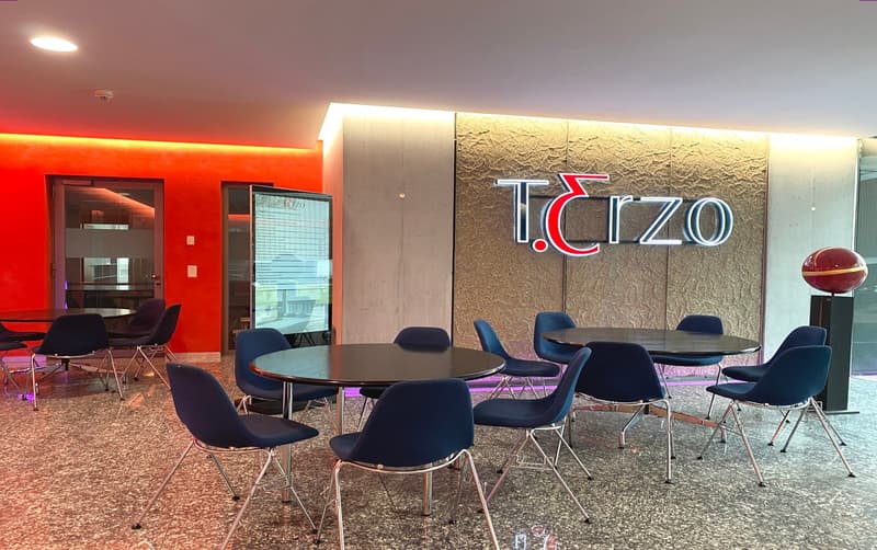 The image shows the interior of an office space belonging to the company Tcizo. The space features a large logo sign on the wall, several round tables with black chairs, and a red and orange color scheme throughout the room.